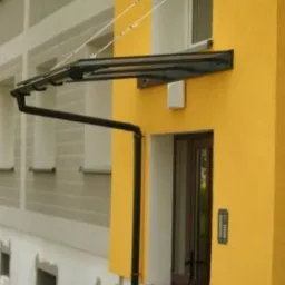 Zadaszenie nad wejściem do budynku z żółtą elewacją, wsparte na stalowych linkach, z widoczną rynną odprowadzającą wodę.