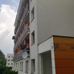 Wielopiętrowy budynek mieszkalny z balkonami, świeżo po malowaniu elewacji na biało, z pomarańczowymi akcentami przy wejściu, widoczne detale takie jak numer budynku i nazwa ulicy, oraz butelki po...