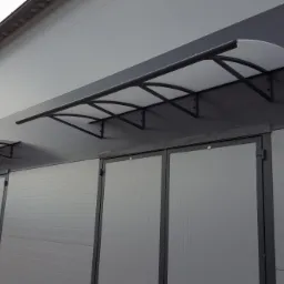 Zadaszenie z poliwęglanu nad dwuskrzydłowymi drzwiami, wsparte na metalowej konstrukcji, zamontowane na szarej elewacji budynku.