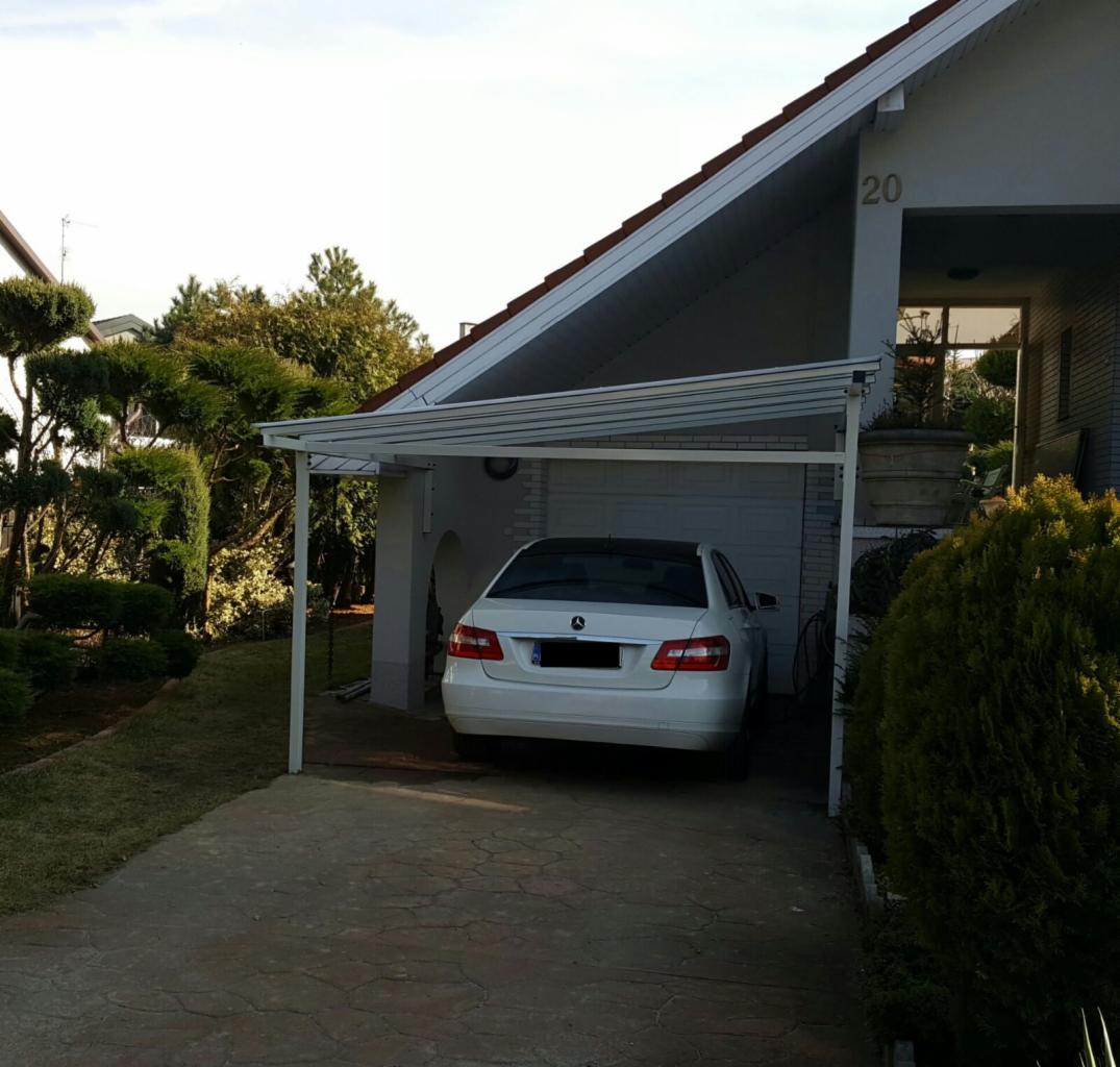 Biały samochód zaparkowany pod nowoczesną, metalową konstrukcją przylegającą do domu z białej cegły, widok z tyłu pojazdu, zadbany trawnik i krzewy w tle.