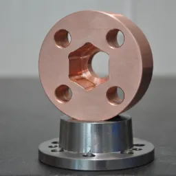 Miedziany element z centralnym sześciokątnym otworem i czterema okrągłymi otworami, umieszczony na srebrnym metalowym cylindrze z kołnierzem, na gładkiej powierzchni.