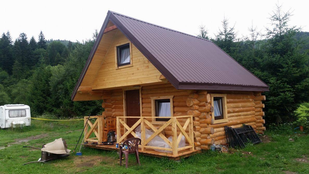 Surowy, drewniany domek z bali z brązowym dachem i nieukończoną werandą, na tle lasu i z przyczepą kempingową w tle.
