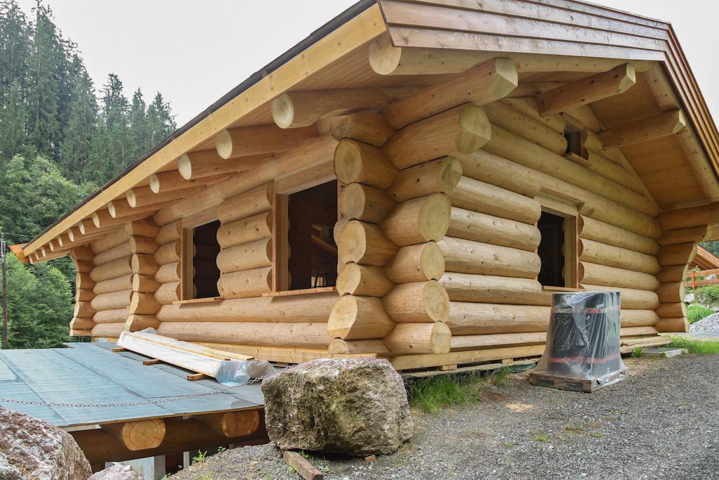 Surowy stan budowy parterowego budynku z bali z widocznymi otworami okiennymi, zabezpieczonymi materiałami budowlanymi i dużym kamieniem na pierwszym planie.