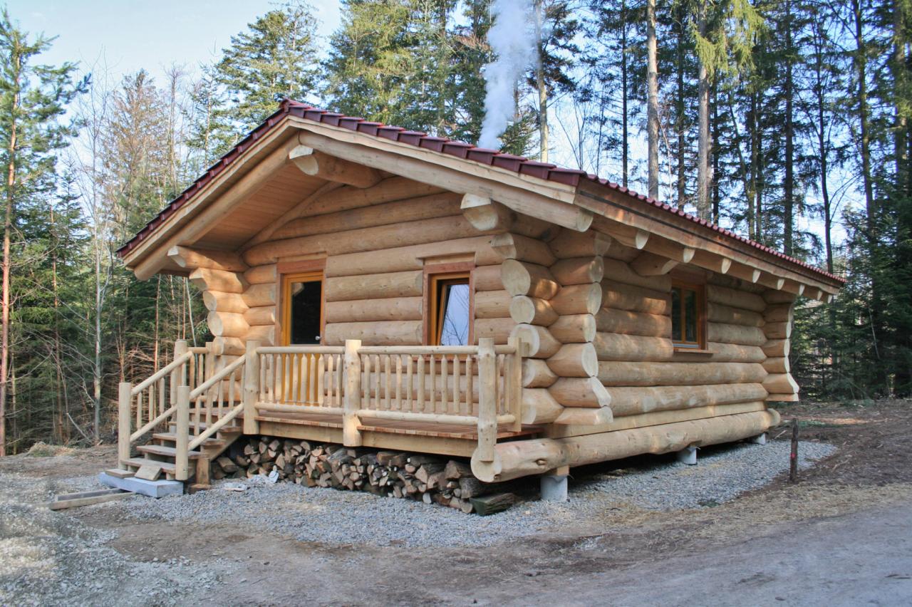 Gotowy, parterowy budynek z bali z czerwonym dachem, widoczny dym z komina i stos drewna pod werandą