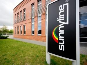 Czarny szyld reklamowy z białym napisem 'sunnyline' i kolorowym łukiem w barwach żółtej, czerwonej i zielonej, na tle ceglanego budynku z trawnikiem