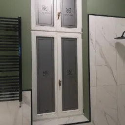 Czterodrzwiowa szafka wnękowa z białymi ramami i szarymi matowymi szybami z czarnym nadrukiem, zamontowana w łazience z zieloną ścianą i marmurowymi płytkami.