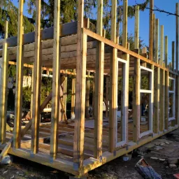 Konstrukcja szkieletowa domu w trakcie budowy, widoczne drewniane ramy, słupy i belki, częściowo osadzone okna, na zewnątrz budynku w tle drzewa i błękitne niebo.