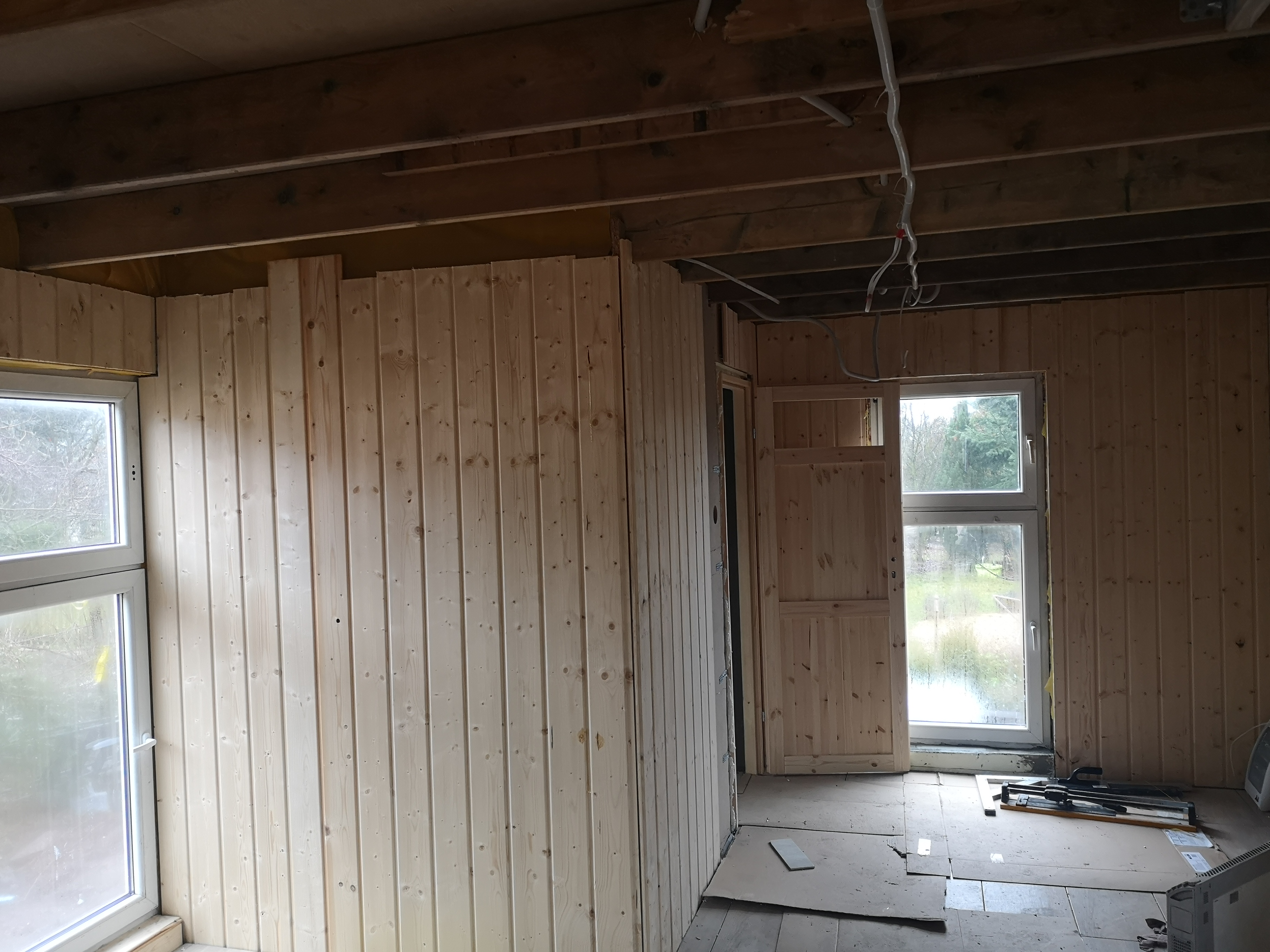 Wnętrze domu szkieletowego w trakcie wykańczania, widoczne drewniane panele ścienne, okna z widokiem na zewnątrz, odsłonięte przewody elektryczne pod sufitem oraz narzędzia na podłodze.