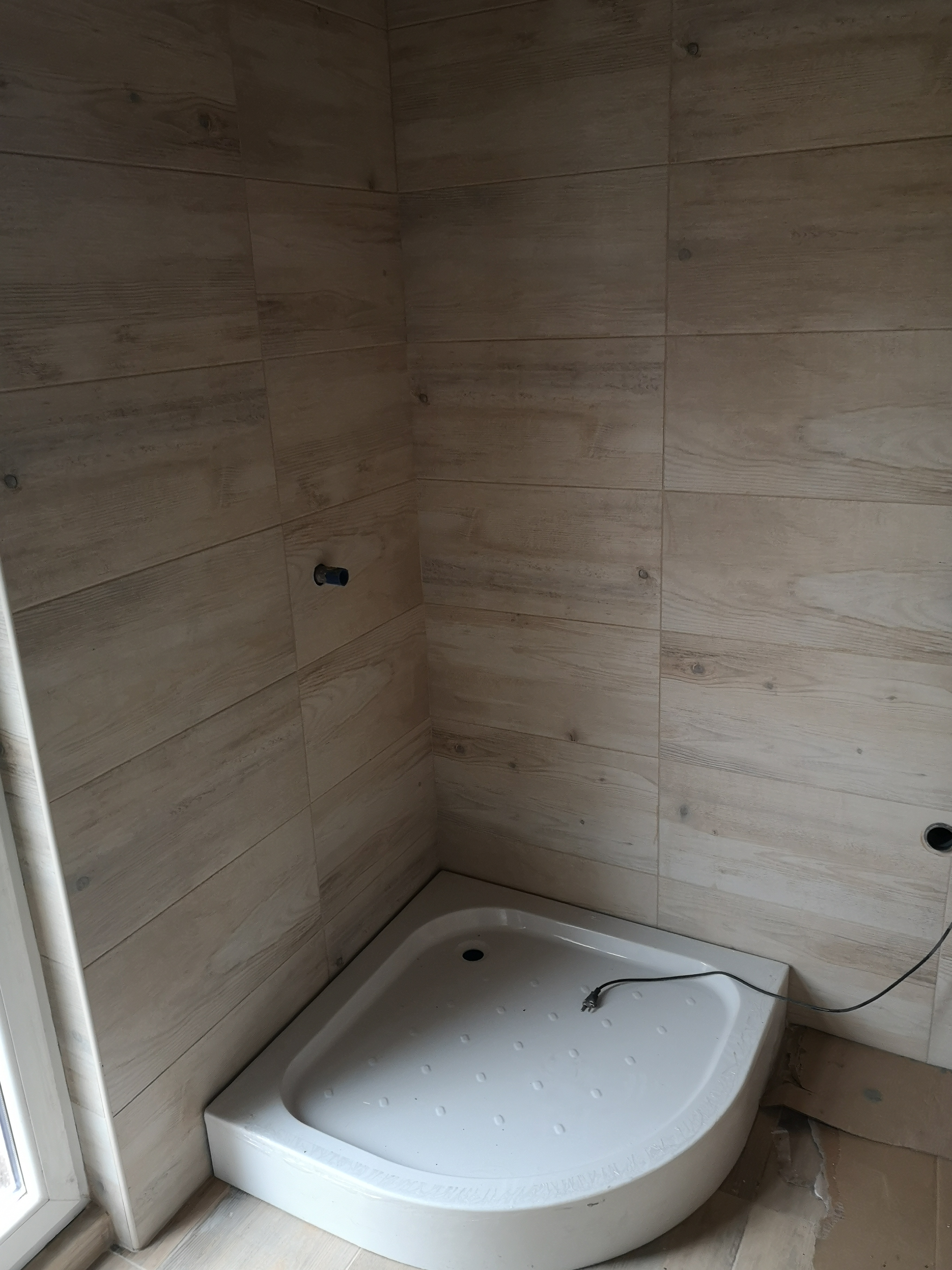 Narożny brodzik z białego akrylu, otoczony płytkami ceramicznymi imitującymi drewno, z widocznym kablem elektrycznym i przygotowanymi otworami na armaturę.