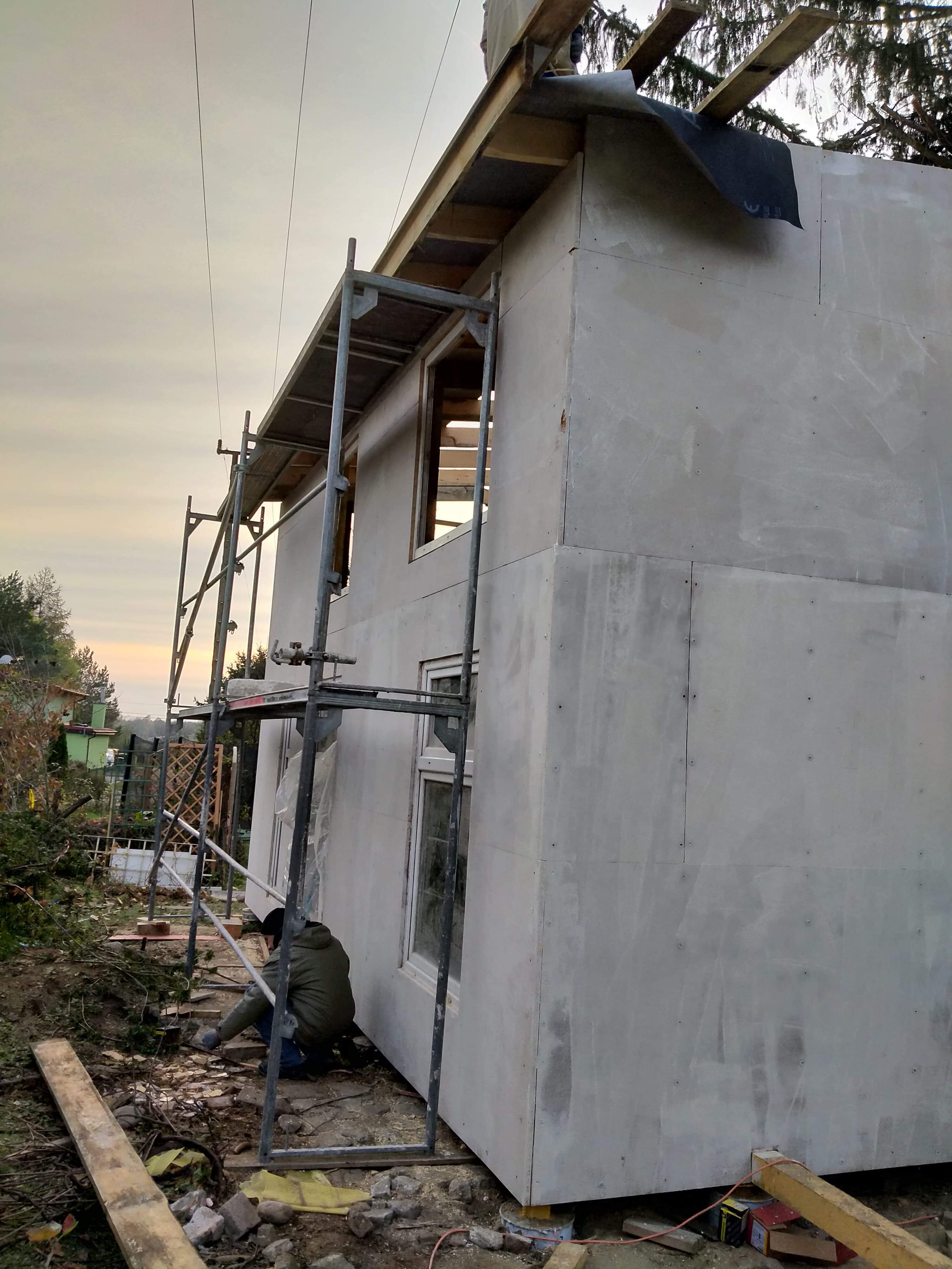 Budowa domu szkieletowego: widok z zewnątrz na częściowo obłożoną płytami ścianę, widoczne rusztowanie i pracownik przy fundamentach.