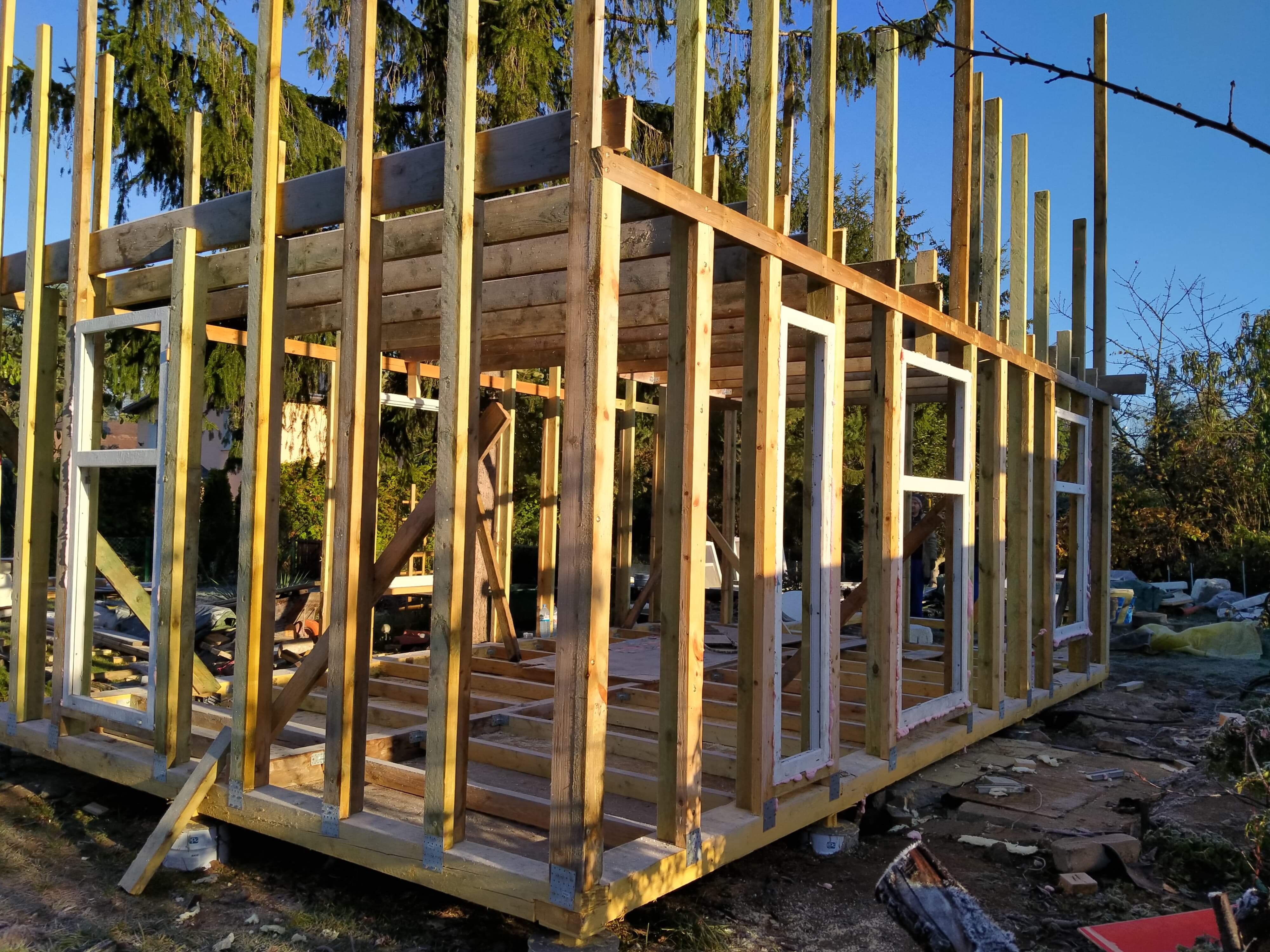 Konstrukcja szkieletowa domu w trakcie budowy, widoczne drewniane ramy, słupy i belki, częściowo osadzone okna, na zewnątrz budynku w tle drzewa i błękitne niebo.