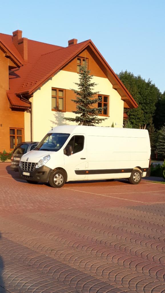 Biały bus Renault Master zaparkowany na kostce brukowej przed budynkiem z pomarańczowym dachem.