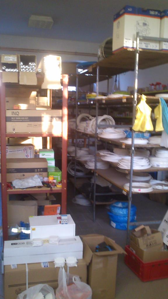 Magazyn z półkami pełnymi zwojów kabli elektrycznych, pudełek z osprzętem, narzędzi i materiałów instalacyjnych, oświetlony naturalnym światłem wpadającym przez okno.