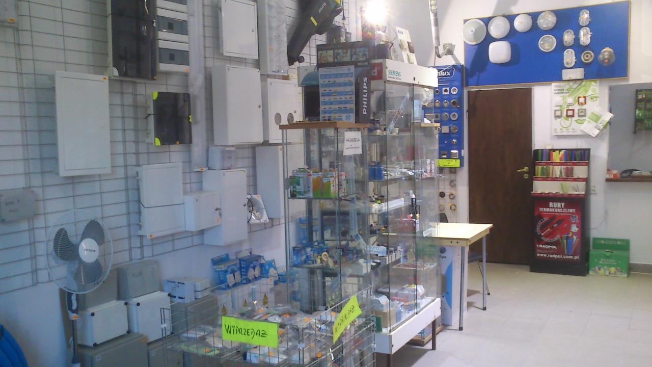 Wnętrze sklepu elektrycznego: ekspozycja z rozdzielnicami na ścianie, szklana witryna z produktami na wyprzedaży, oświetlenie LED na niebieskiej tablicy i materiały instalacyjne.