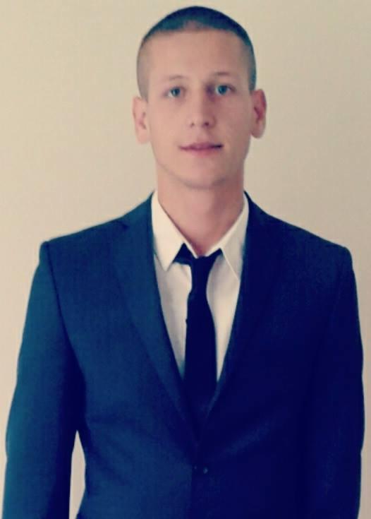 Portret młodego mężczyzny w ciemnym garniturze i białej koszuli z czarnym krawatem, stojącego na jasnym tle.