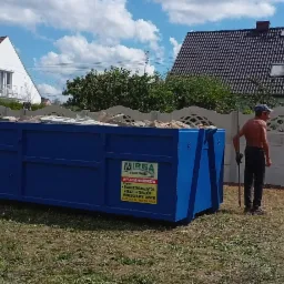 Niebieski kontener wypełniony gruzem stoi na trawniku przed domem; mężczyzna bez koszulki opiera się o łopatę obok kontenera, w tle widoczny betonowy płot i dwa domy.