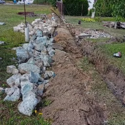 Wykopany rów w ziemi, obok sterta gruzu z niebieskawym odcieniem, widoczne narzędzia i elementy konstrukcyjne w tle, krajobraz z domami.