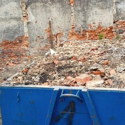 Etna - Niebieski kontener wypełniony gruzem ceglanym i betonowym na tle ściany budynku w trakcie rozbiórki, z widocznymi fragmentami cegieł i resztkami tynku.