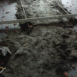 Etna - Wylewka betonowa na podłodze z rozłożoną siatką zbrojeniową i poziomnicą, widoczne ślady butów w betonie, fragment ściany w tle.