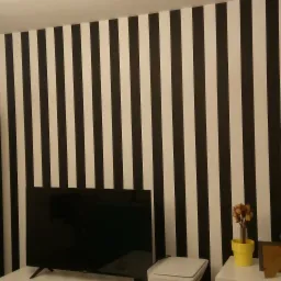 Pokój z biało-czarną tapetą w pionowe pasy, białą szafką z przeszklonymi drzwiami, telewizorem, kwiatami w doniczkach i dekoracjami na białej komodzie.