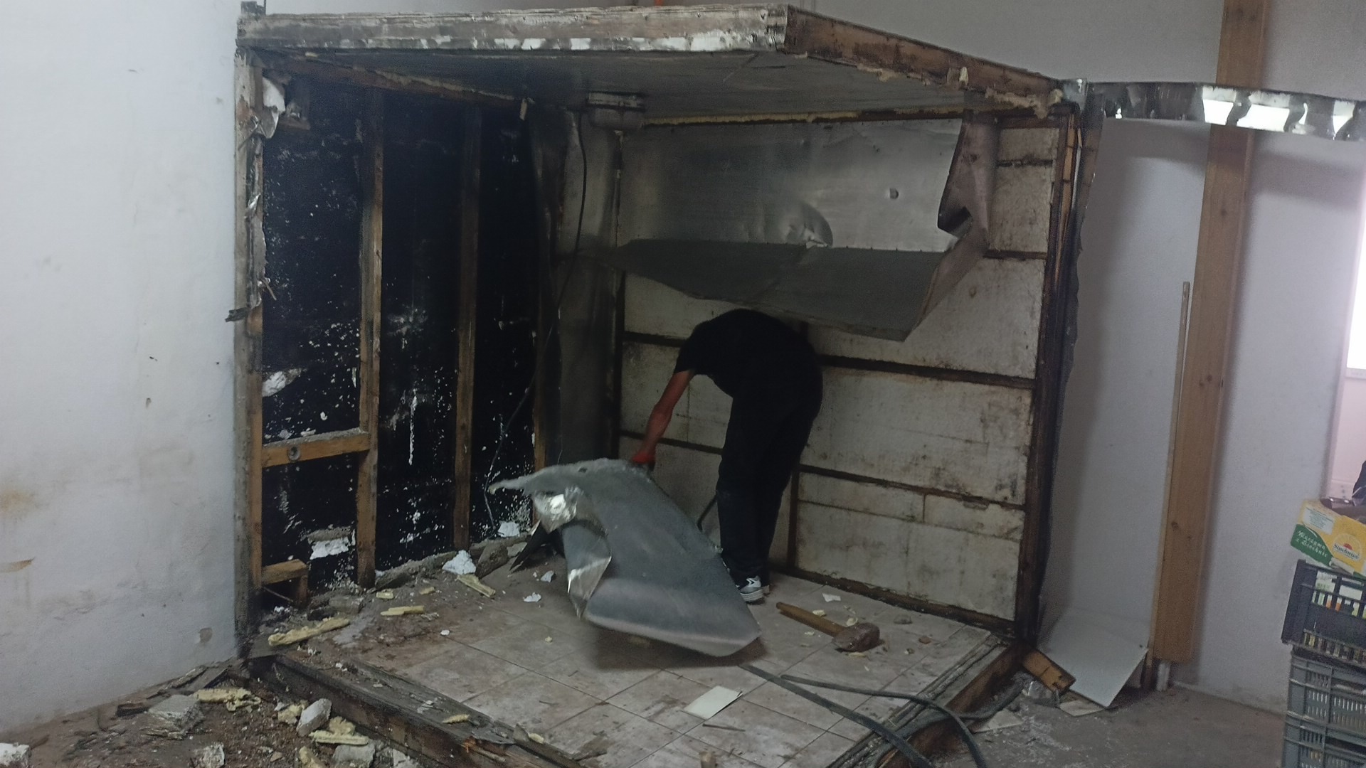 Demontaż konstrukcji wewnątrz pomieszczenia: mężczyzna w czarnym ubraniu usuwa metalowy element, widoczne pozostałości ścian, podłoga pokryta gruzem i fragmentami materiałów budowlanych.