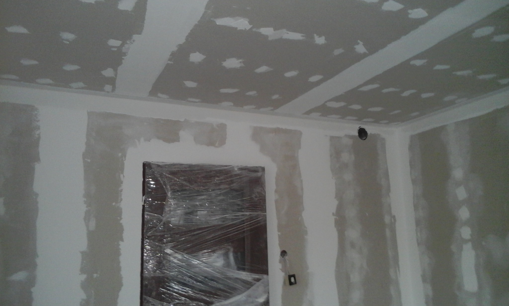 Ściany i sufit w trakcie szpachlowania, zabezpieczone folią drzwi w tle, widoczne puszki elektryczne.