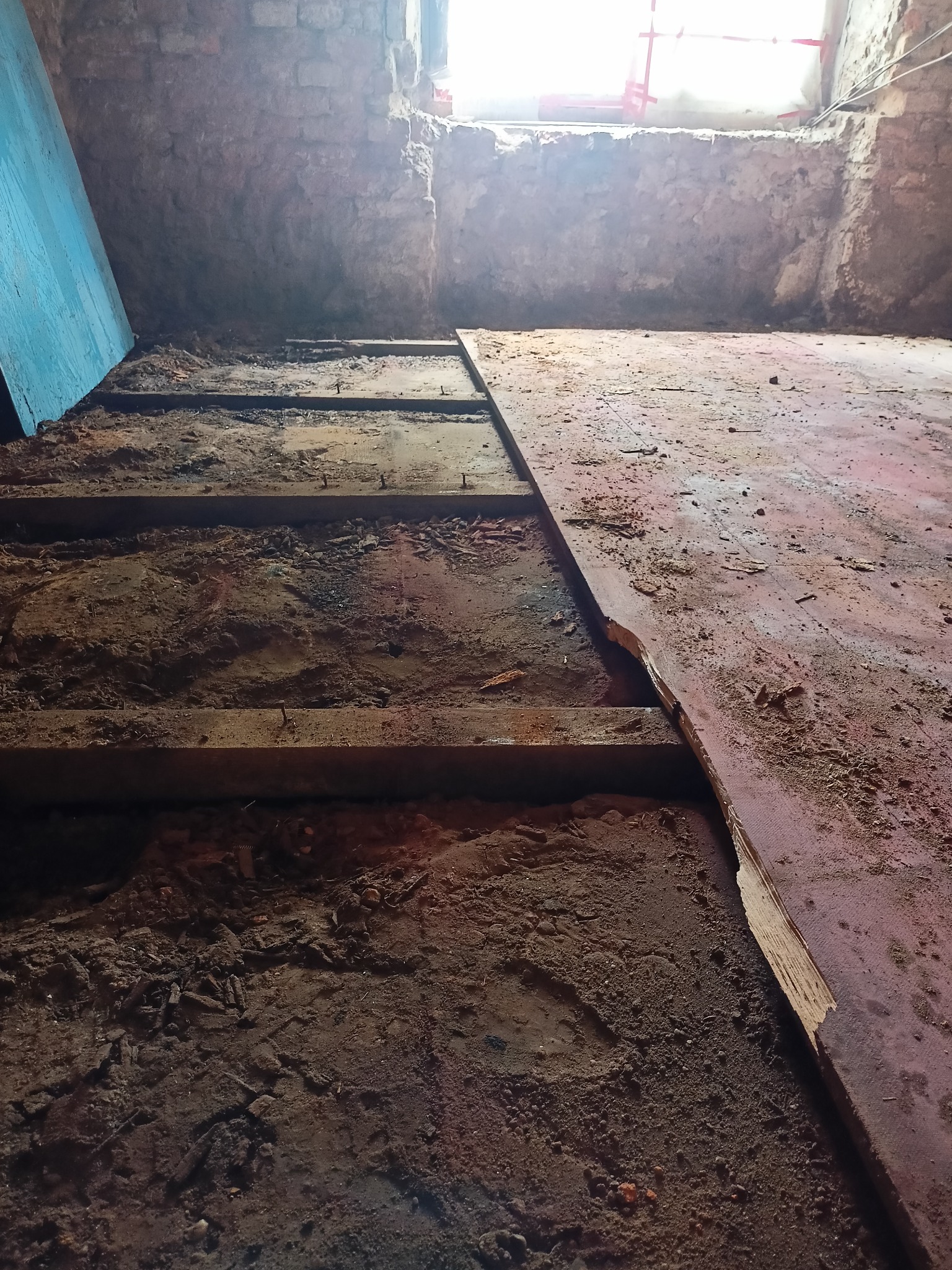 Demontaż podłogi w piwnicy ze starą cegłą, widoczne deski podłogowe i gruz, w tle okno z rozproszonym światłem.