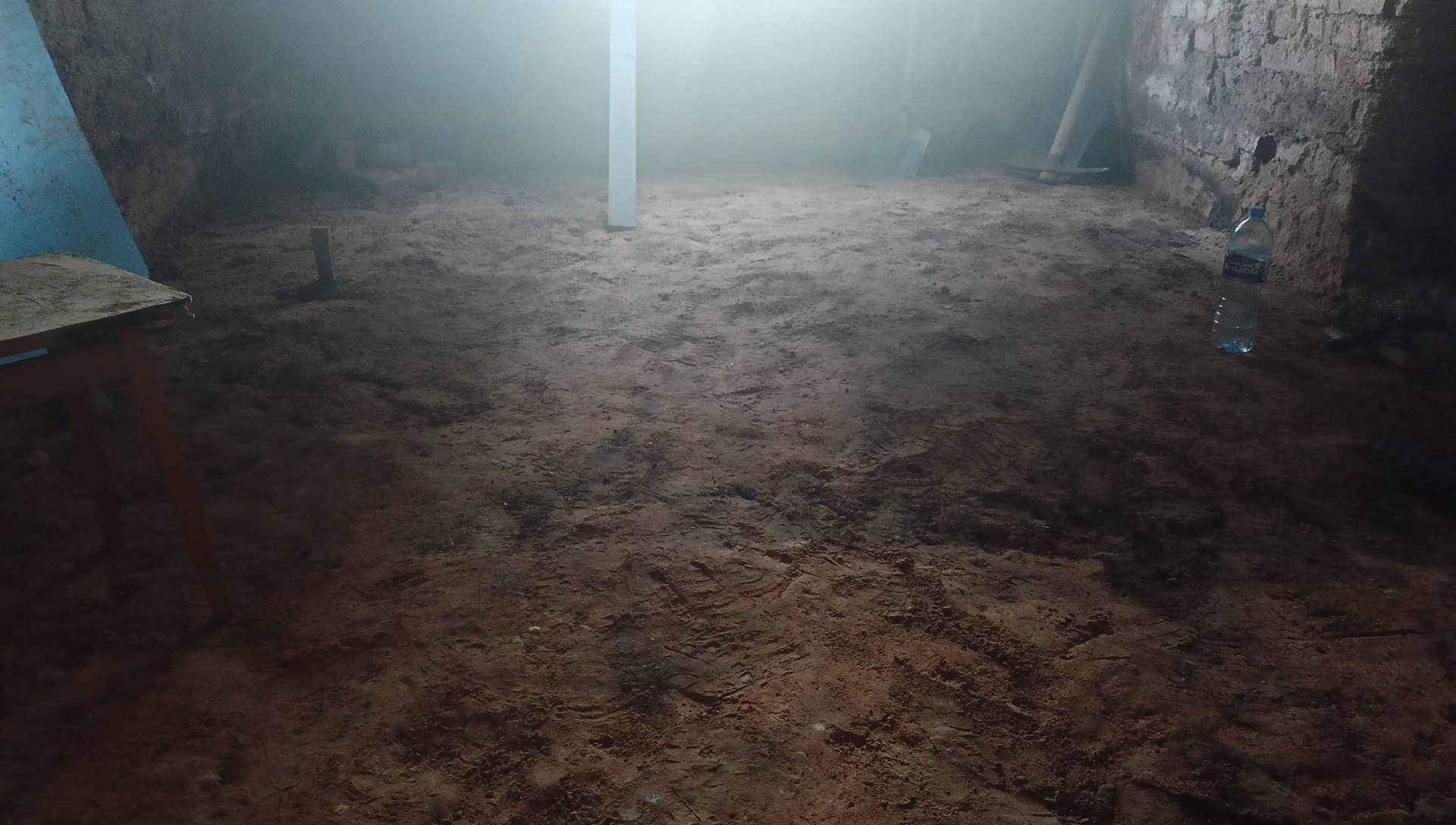 Przygotowany grunt pod wylewkę w słabo oświetlonym pomieszczeniu, widoczne ślady stóp, narzędzia i butelka wody.
