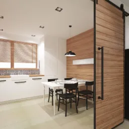 Nowoczesna kuchnia z białymi szafkami, drewnianym blatem i wzorzystą glazurą nad blatem, uzupełniona o stół z czarnymi krzesłami i przesuwanymi drzwiami z desek oraz kominek z drewnem.