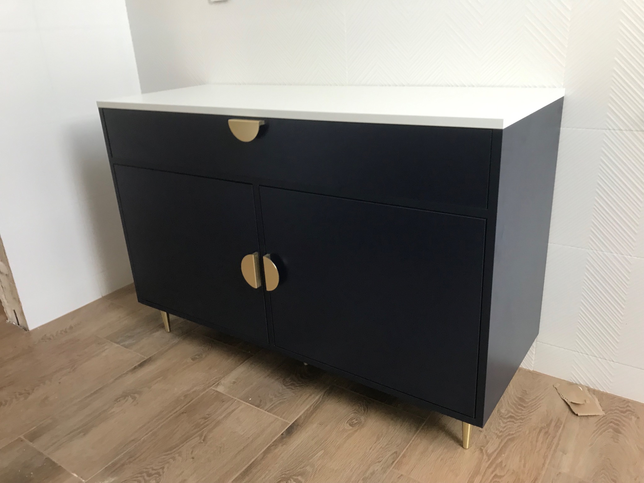 Granatowa szafka z białym blatem i złotymi uchwytami na cienkich nóżkach, ustawiona na drewnopodobnej podłodze w pomieszczeniu z białą, wzorzystą ścianą.