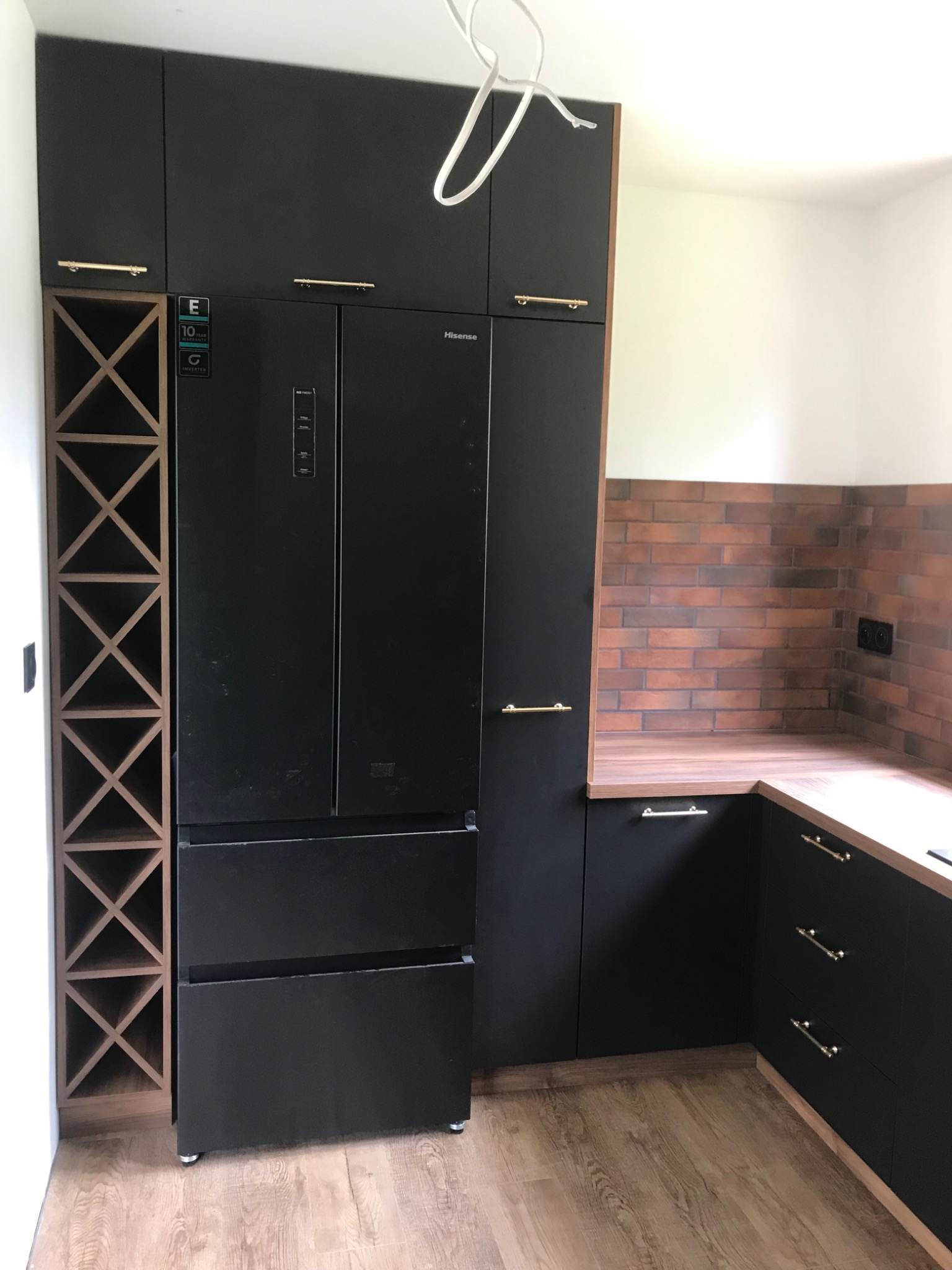 Czarna zabudowa kuchenna z lodówką typu side-by-side i drewnianym regałem na wino, blat z imitacji drewna, ściana wyłożona cegłą.