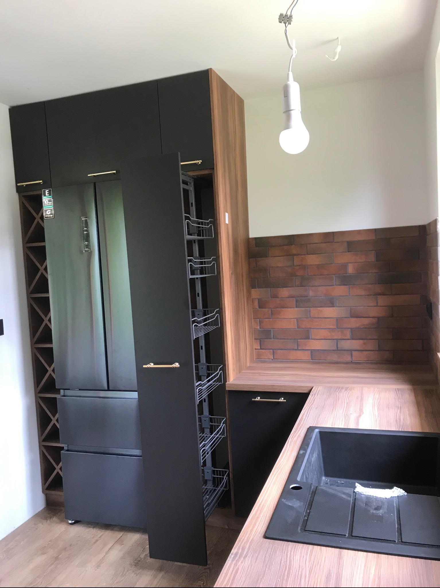 Czarna zabudowa kuchenna z wysuwaną szafką cargo, lodówką side-by-side i regałem na wino, uzupełniona blatem imitującym drewno i ceglaną ścianą.