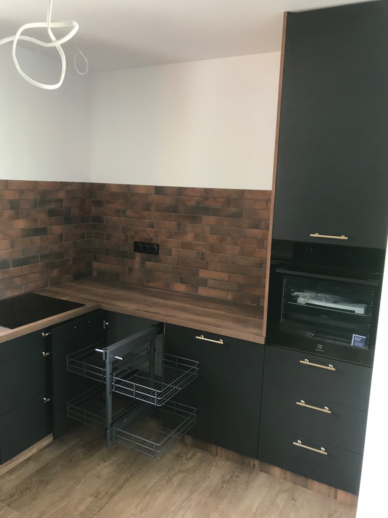 Nowoczesna kuchnia z czarnymi frontami, drewnianym blatem i ceglaną ścianą, wysuwany kosz cargo w szafce narożnej.