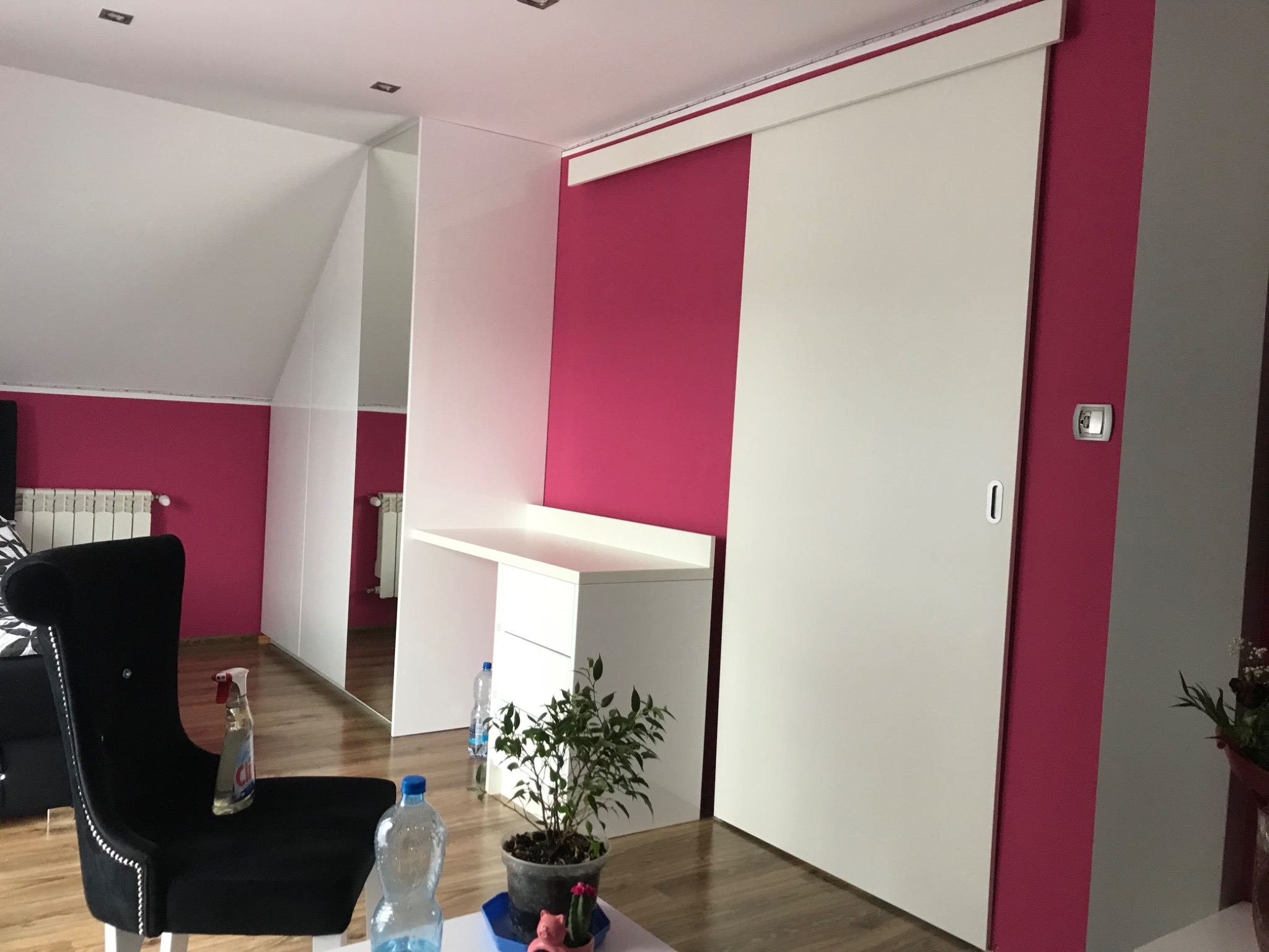 Biała szafa wnękowa z przesuwnymi drzwiami w pokoju na poddaszu w Zbytkowie, z różowymi ścianami i czarnym fotelem w stylu glamour.
