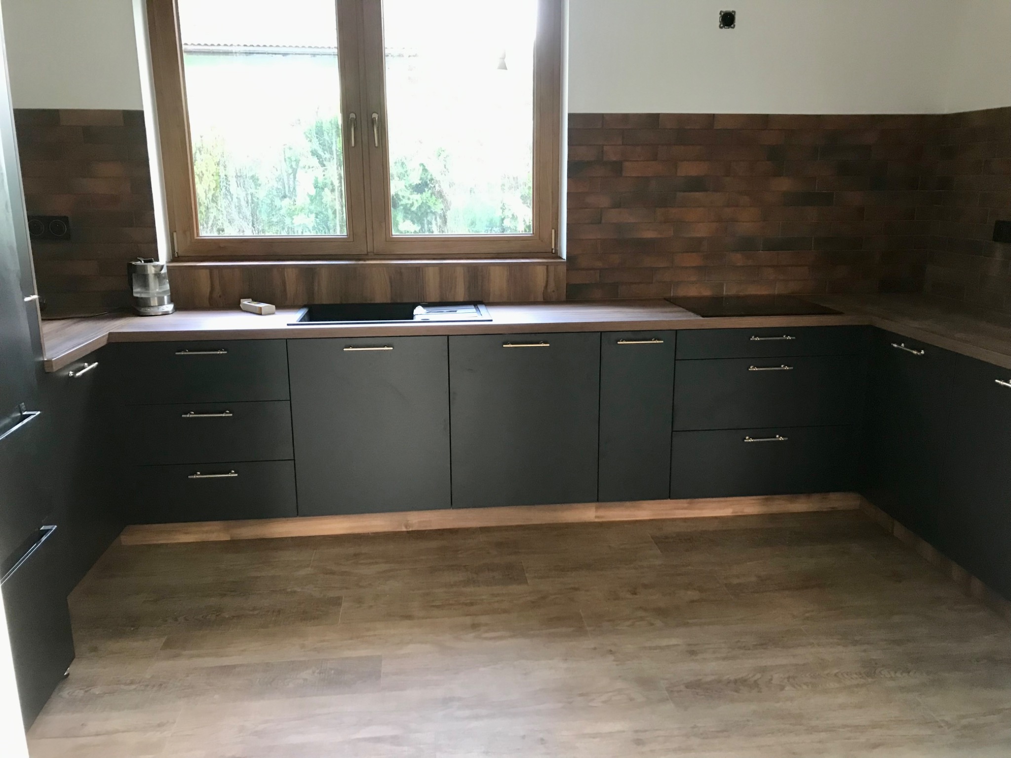 Nowoczesna kuchnia z ciemnymi, matowymi frontami szafek, drewnianym blatem i ceglaną ścianą nad blatem, widok na okno z zewnątrz