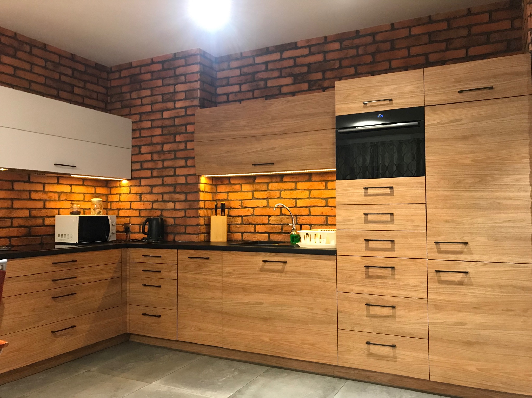 Jasna, drewniana zabudowa kuchenna z czarnym blatem i ceglaną ścianą w tle, oświetlona ciepłym światłem.