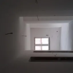 Wnętrze w trakcie prac wykończeniowych; widoczne ściany po nałożeniu pierwszej warstwy, okno w tle, wystające kable i niedokończone obramowanie.