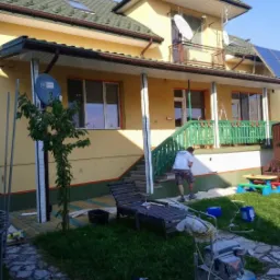 Prace przy elewacji domu jednorodzinnego z zielonym dachem, widoczny agregat malarski i osoba w trakcie natryskowego nakładania farby na dolną część ściany.