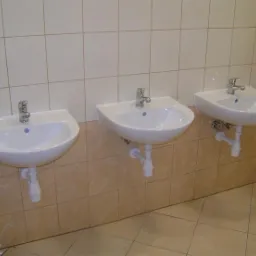 Trzy identyczne białe umywalki zamontowane na ścianie z beżowych płytek, każda z chromowaną baterią i białym syfonem, widoczne rury odpływowe.