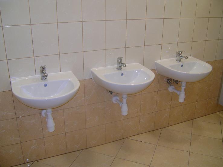 Trzy identyczne białe umywalki zamontowane na ścianie z beżowych płytek, każda z chromowaną baterią i białym syfonem, widoczne rury odpływowe.