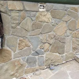 Ściana zewnętrzna wykończona nieregularnymi kamieniami w odcieniach beżu i szarości, z widocznym fragmentem okna, elementami roślinności oraz ozdobnym naczyniem u podstawy ściany.