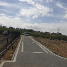 Ułożona kostka brukowa w kolorze szarym z białymi obramowaniami miejsc parkingowych, widok z poziomu gruntu, w tle zielone wzgórza i błękitne niebo z chmurami oraz zaparkowany biały bus.