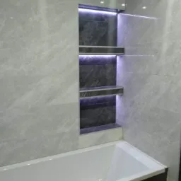 Wykończona łazienka z wanną i wnęką z półkami, podświetloną fioletowym światłem LED, obłożona szarymi płytkami imitującymi marmur.