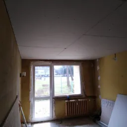 Montaż sufitu z płyt kartonowo-gipsowych w trakcie remontu mieszkania w Zakopanem, widoczne płyty przymocowane do konstrukcji, okno z widokiem na zewnątrz, grzejnik i częściowo przygotowane ściany.