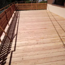 Nowo wybudowany drewniany taras z jasnego drewna, otoczony niskim płotkiem, z widocznymi cieniami barierki, na tle drzew i budynku.