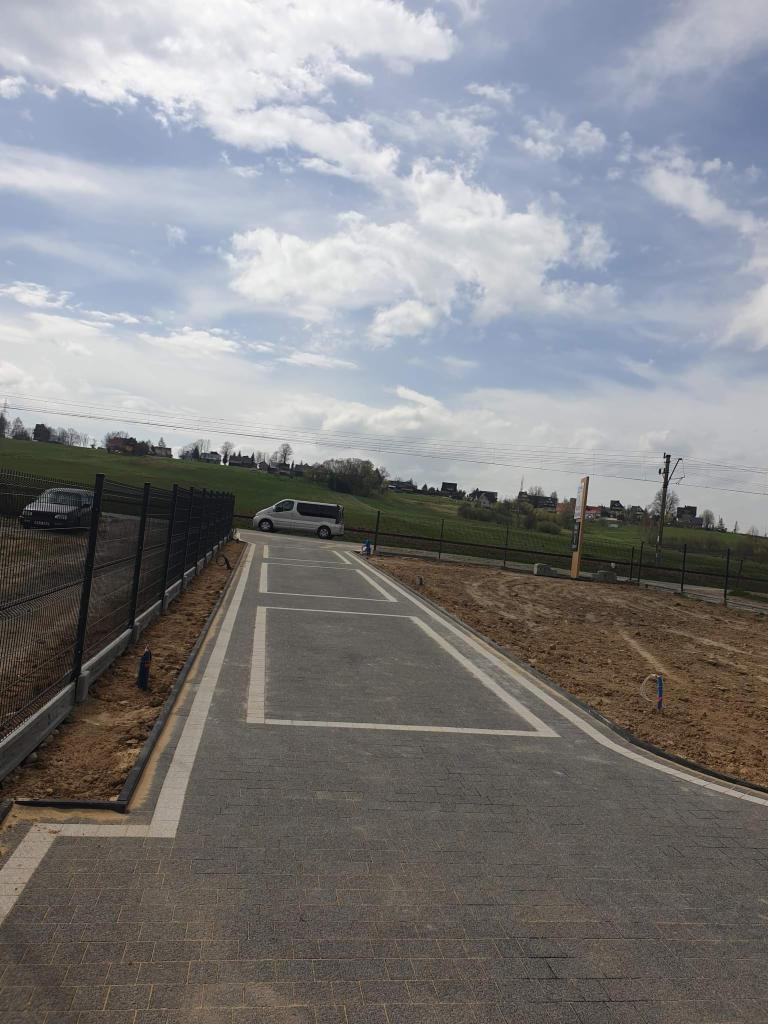 Ułożona kostka brukowa w kolorze szarym z białymi obramowaniami miejsc parkingowych, widok z poziomu gruntu, w tle zielone wzgórza i błękitne niebo z chmurami oraz zaparkowany biały bus.