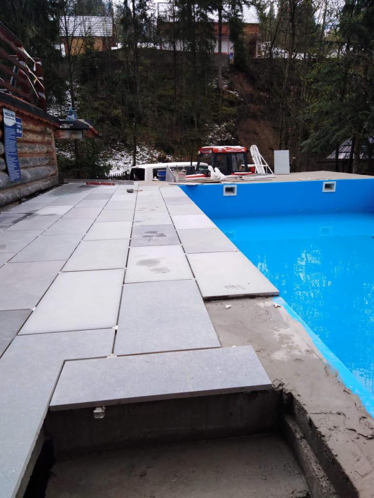 Układanie szarych płyt chodnikowych wokół basenu w Zakopanem, widoczny fragment basenu z błękitną wodą, w tle budynek z bali i zalesione wzgórze.
