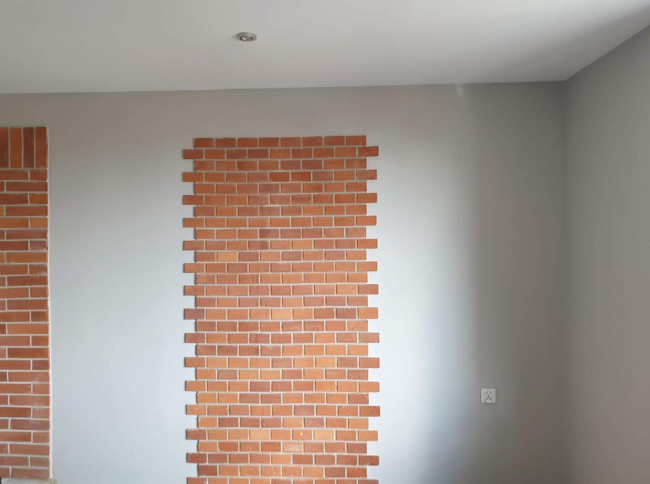 Fragment ściany wewnętrznej z częściowo ułożoną dekoracyjną cegłą w odcieniach czerwieni i pomarańczy na tle szarej ściany, widoczne niedokończone pasy cegieł.