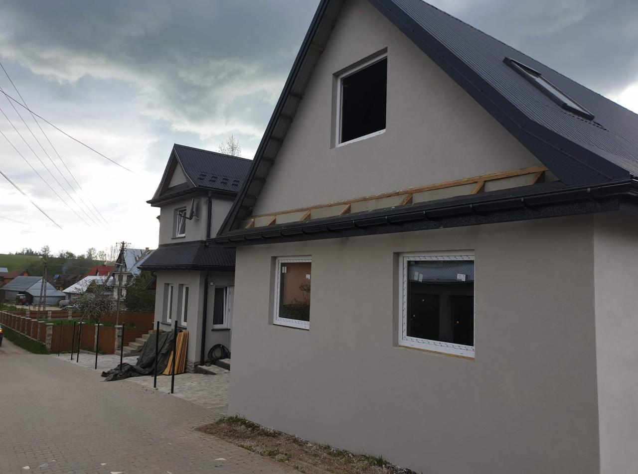 Szary dom jednorodzinny z czarnym dachem w trakcie remontu, widoczne nowe okna i przygotowanie do montażu podbitki dachowej, pochmurne niebo w tle.