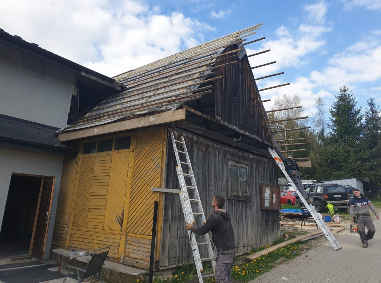 Dwóch mężczyzn na drabinach podczas remontu dachu starego, drewnianego budynku z żółtą elewacją, widoczne krokwie i membrana dachowa, w tle drzewa i samochody.